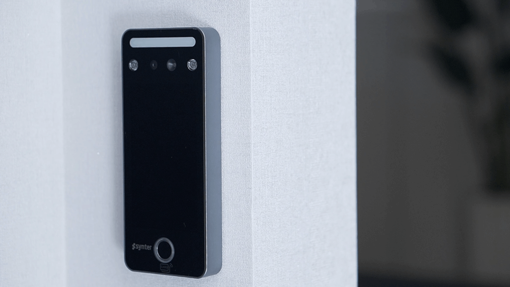 Interfone digital Synter instalado em parede branca, com design moderno e minimalista, mostrando câmera, sensores e botão de chamada